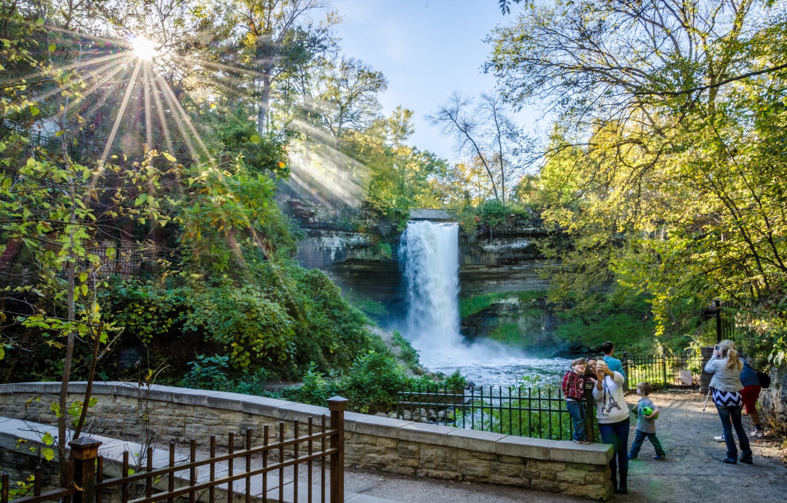 Minnehaha Falls in Minneapolis with sunlight streaming through the trees and visitors gathered at the overlook railing below.