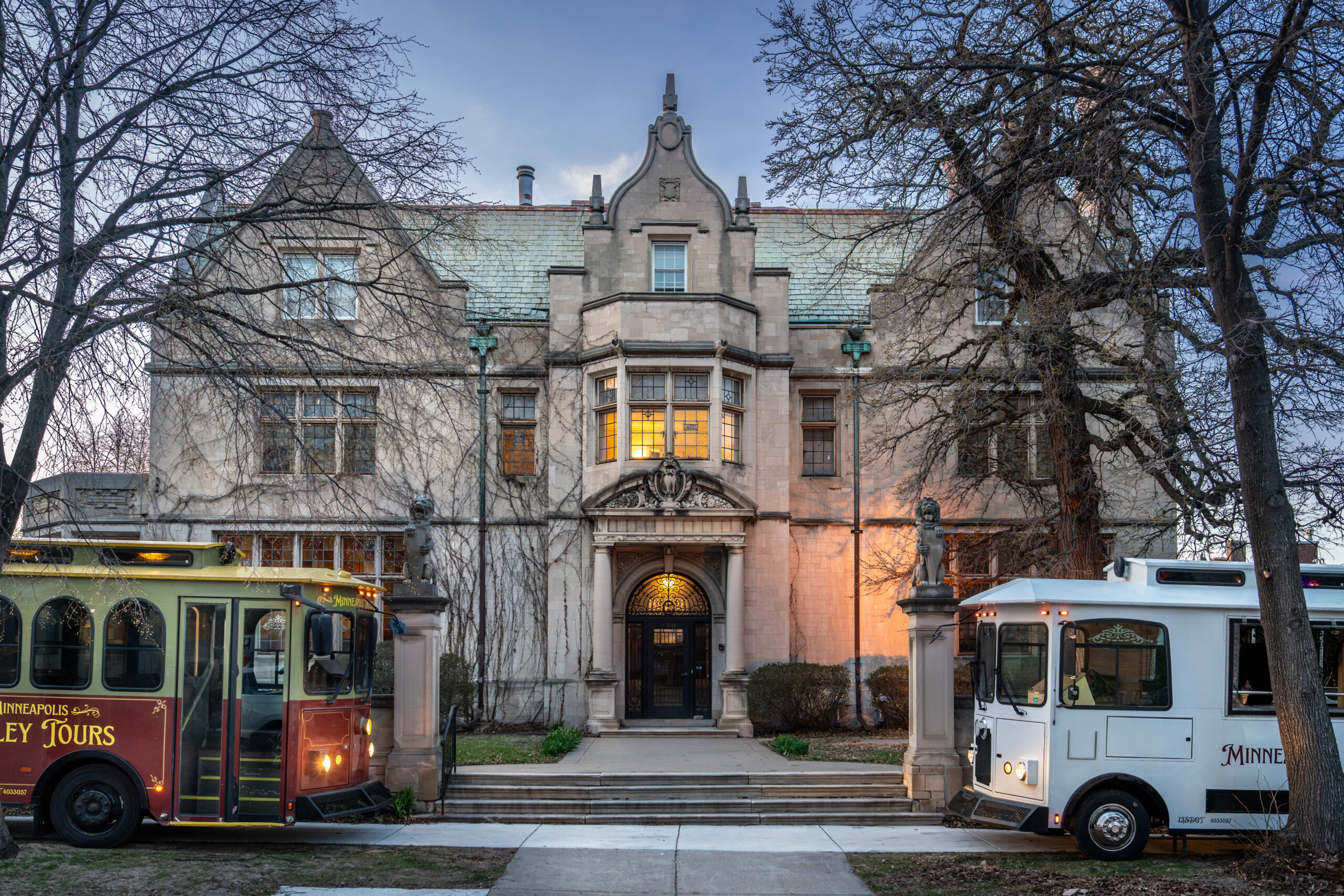 Two Minneapolis Trolley Tours vehicles parked in front of the historic Pillsbury Mansion at dusk, the departure point for sightseeing, ghost and Gilded Age mansion tours in Minneapolis